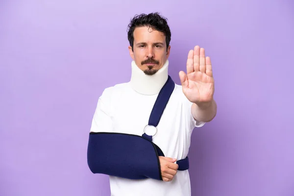 stock image Young caucasian man wearing a sling and neck brace isolated on purple background making stop gesture