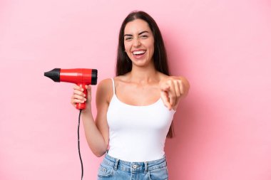 Pembe arka planda izole edilmiş saç kurutma makinesini tutan beyaz bir kadın kendine güvenen bir ifadeyle sana parmak kaldırıyor.