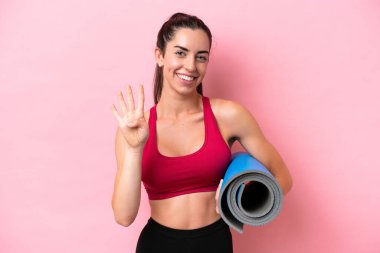 Genç beyaz kadın yoga derslerine gidiyor pembe arka planda izole edilmiş bir minderi tutarken mutlu ve parmaklarıyla dörde kadar sayıyor.