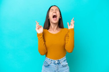 Mavi arka planda izole edilmiş genç beyaz kadın parmaklarını çapraz yapıp en iyisini diliyor.