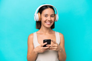 Kafkasyalı genç bir kadın mavi arka planda izole edilmiş. Cep telefonuyla müzik dinliyor ve ön tarafa bakıyor.