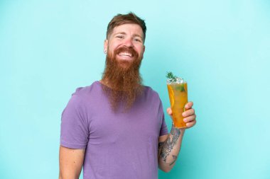 Uzun sakallı kızıl saçlı bir adam mavi arka planda izole edilmiş bir kokteyl tutuyor ve gülümsüyor.