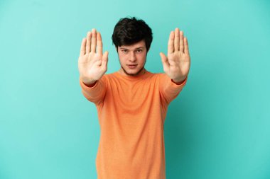 Genç Rus adam mavi arka planda izole edilmiş. Durma hareketi yapıyor ve hayal kırıklığına uğruyor.