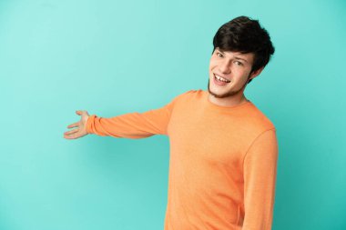 Genç Rus adam mavi arka planda izole edilmiş. Davet etmek için ellerini yana uzatıyor.