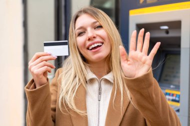 Genç, güzel, sarışın bir kadın dışarıda elinde bir kredi kartıyla elini sallayarak mutlu bir ifadeyle selam veriyor.
