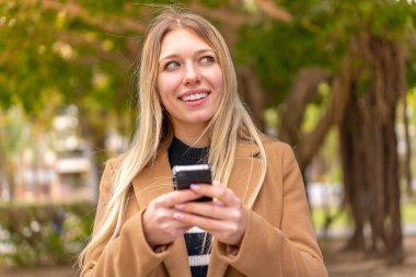 Dışarıdaki güzel sarışın kadın cep telefonu kullanıyor ve yukarı bakıyor.