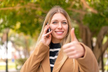 Açık havada genç, güzel sarışın bir kadın başparmak kaldırırken cep telefonuyla sohbet ediyor.