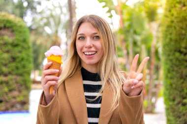 Açık havada kornet dondurmalı güzel sarışın kadın gülümsüyor ve zafer işareti gösteriyor.