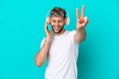 Genç yakışıklı beyaz adam mavi arka planda izole edilmiş müzik dinliyor ve şarkı söylüyor.