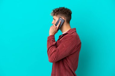 Mavi arka planda izole edilmiş yakışıklı beyaz bir adam cep telefonuyla sohbet ediyor.