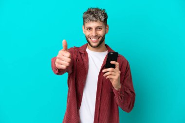 Young handsome caucasian man isolated on blue background using mobile phone while doing thumbs up