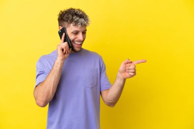 Sarı arka planda izole edilmiş cep telefonu kullanan genç, beyaz bir adam parmak göstererek bir ürün sunuyor.