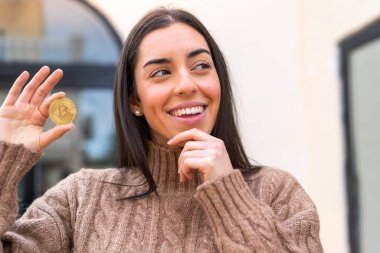 Elinde Bitcoin tutan genç bir kadın bir fikir üretiyor ve ona bakıyor.