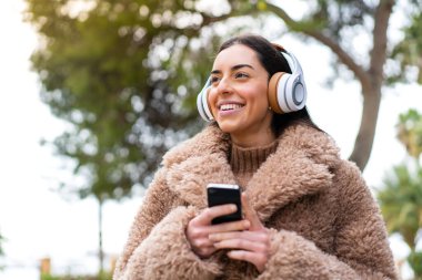 Kadın açık havada cep telefonuyla müzik dinliyor.