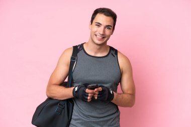 Spor çantalı genç beyaz adam pembe arka planda izole edilmiş cep telefonuyla bir mesaj gönderiyor.