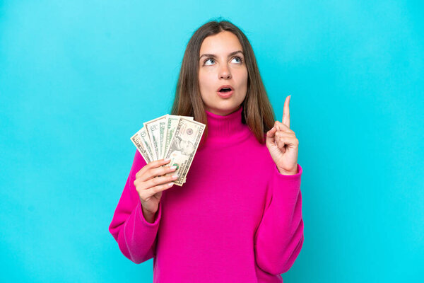 Young caucasian woman taking a lot of money isolated on blue background thinking an idea pointing the finger up