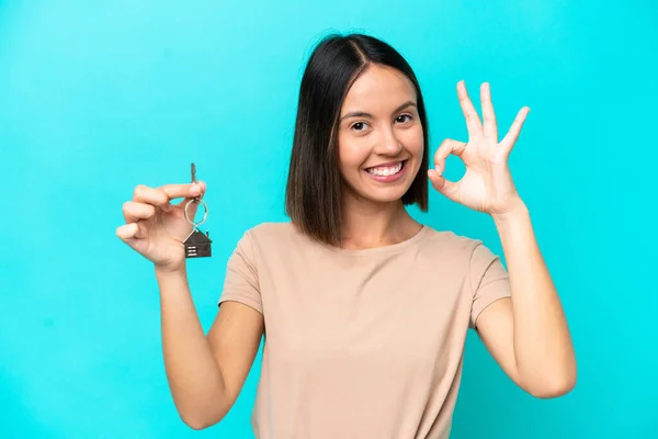 年轻女人拿着家里的钥匙 蓝色背景隔离 用手指显示好的标志 — 图库照片