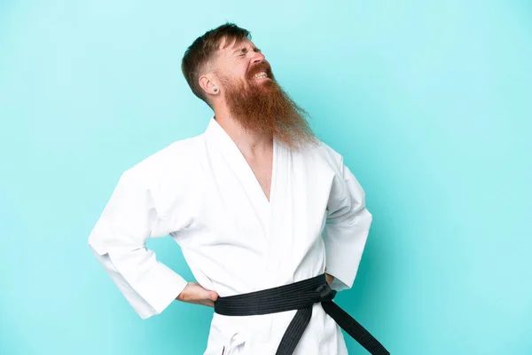 Redhead man with long beard doing karate isolated on blue background suffering from backache for having made an effort
