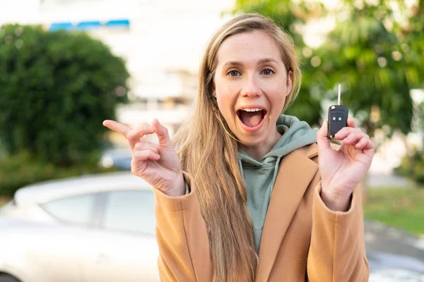 Young blonde woman holding car keys at outdoors surprised and pointing finger to the side