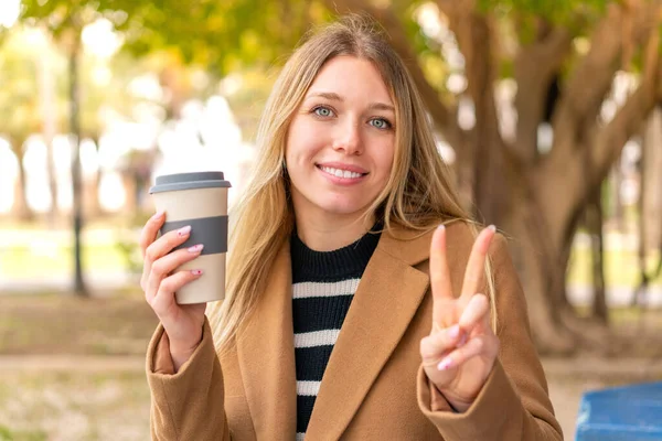 年轻漂亮的金发女子在户外端着一杯咖啡 面带微笑 并显示出胜利的迹象 — 图库照片