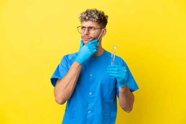 Dentysta Kaukaski Mężczyzna Trzyma Narzędzia Izolowane Żółtym Tle Mając Wątpliwości — Zdjęcie stockowe