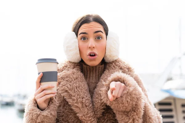 年轻女人穿着冬衣 在户外拿着咖啡 惊讶地指着前面 — 图库照片