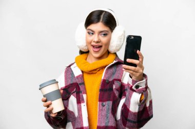 Young Russian woman wearing winter muffs over isolated white background holding coffee to take away and a mobile clipart