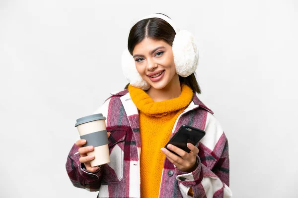 stock image Young Russian woman wearing winter muffs over isolated white background holding coffee to take away and a mobile