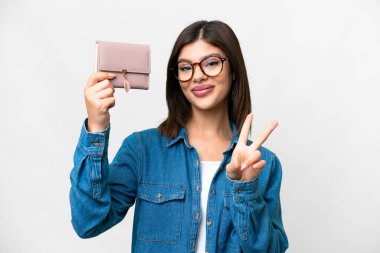 İzole edilmiş beyaz arka planda cüzdanını tutan genç Rus kadın gülümsüyor ve zafer işareti gösteriyor.