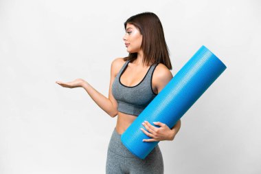 Genç sporcu kadın yoga derslerine gidiyor. Elinde paspas tutarken beyaz bir zemin var. Sürpriz bir ifadeyle yan tarafa bakıyor.