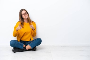 Beyaz arka planda izole bir şekilde yerde oturan beyaz bir kadın ön tarafı işaret ediyor ve gülümsüyor.