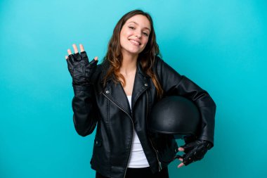 Young caucasian woman with a motorcycle helmet isolated on blue background saluting with hand with happy expression