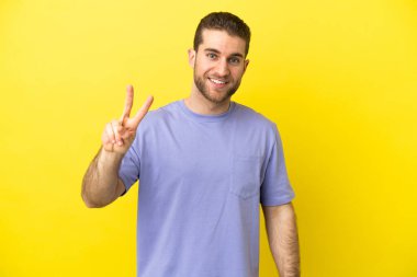 Sarışın yakışıklı adam izole edilmiş sarı arka planda gülümsüyor ve zafer işareti gösteriyor.
