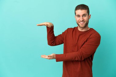 İzole edilmiş mavi arkaplan üzerinde yakışıklı sarışın bir adam bir reklam eklemek için kopyalama alanı tutuyor