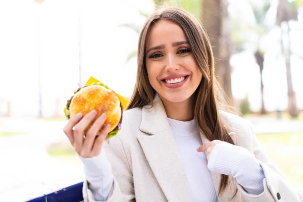 Młoda Ładna Kobieta Trzyma Hamburgera Świeżym Powietrzu Zaskakującym Wyrazem Twarzy — Zdjęcie stockowe