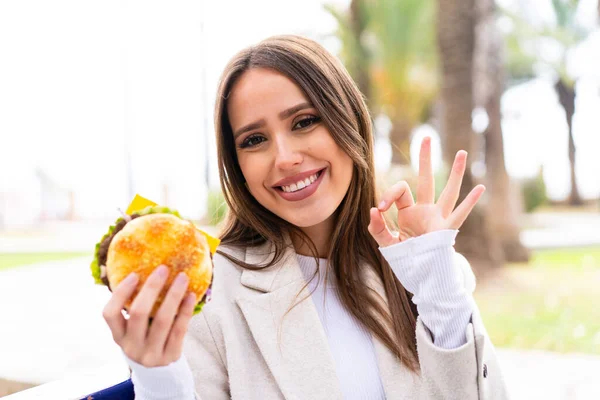 Młoda Ładna Kobieta Trzyma Hamburgera Świeżym Powietrzu Pokazując Znak Palcami — Zdjęcie stockowe