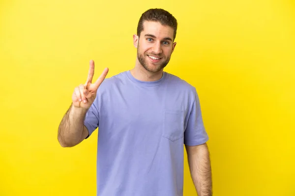 ハンサムなブロンド男上の隔離された黄色の背景笑顔とショー勝利サイン — ストック写真