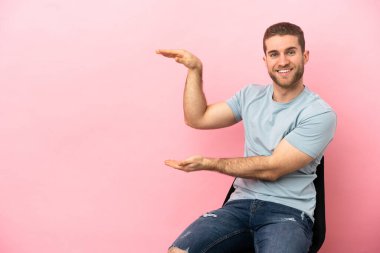 İzole edilmiş pembe arka planda bir sandalyede oturan genç adam reklam koymak için kopyalama alanı tutuyor.