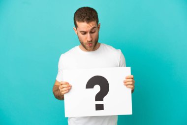 İzole edilmiş mavi arkaplan üzerinde soru işareti olan bir pankart tutan yakışıklı sarışın adam.