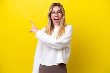 Sarı arka planda izole edilmiş genç Uruguaylı kadın şaşırmış ve işaret ediyor.