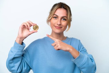 Genç Uruguaylı kadın elinde Bitcoin ile beyaz arka planda izole edilmiş bir şekilde bir ürün sunmak için kenarı işaret ediyor.