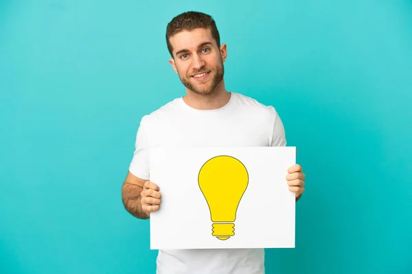 stock image Handsome blonde man over isolated blue background holding a placard with bulb icon with happy expression