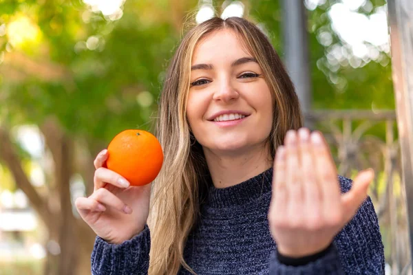 Young pretty Romanian woman holding an orange at outdoors inviting to come with hand. Happy that you came