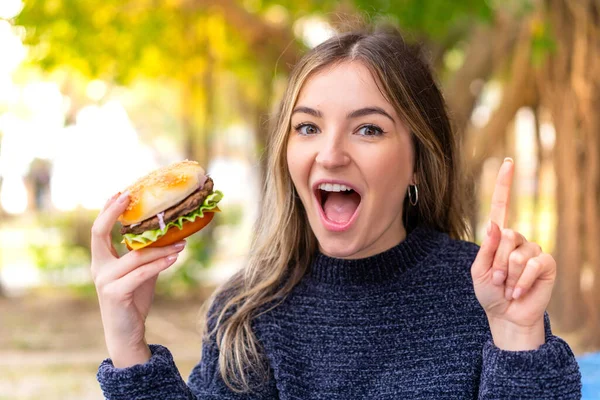 Młoda Ładna Rumuńska Kobieta Trzyma Hamburgera Świeżym Powietrzu Wskazując Świetny — Zdjęcie stockowe