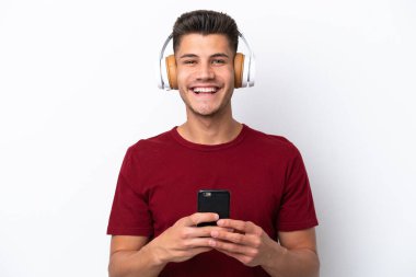 Beyaz arka planda izole edilmiş genç beyaz bir adam cep telefonuyla müzik dinliyor ve ön tarafa bakıyor.