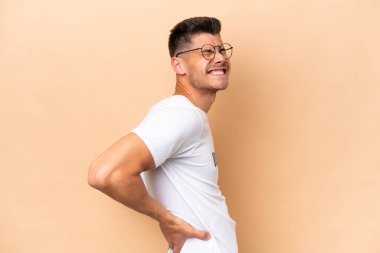 Genç, beyaz, bej arka planda izole edilmiş bir adam gayret gösterdiği için sırt ağrısı çekiyor.