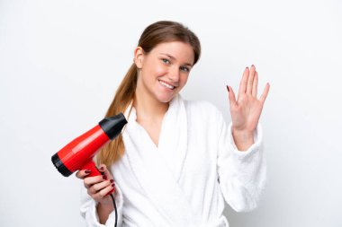 Beyaz arka planda elinde saç kurutma makinesi tutan beyaz bir kadın mutlu bir ifadeyle selam veriyor.