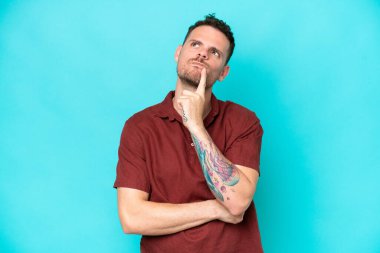 Kafkasyalı yakışıklı genç adam mavi arka planda izole edilmiş, yukarı bakarken şüpheleri var.