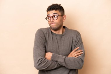 Genç Brezilyalı adam bej arka planda izole edilmiş. Omuzlarını kaldırırken şüpheli hareketler yapıyor.