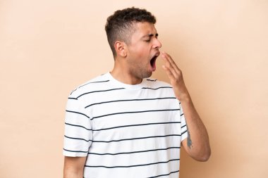 Genç Brezilyalı bir adam bej arka planda izole edilmiş esniyor ve geniş bir ağzı eliyle kapatıyor.